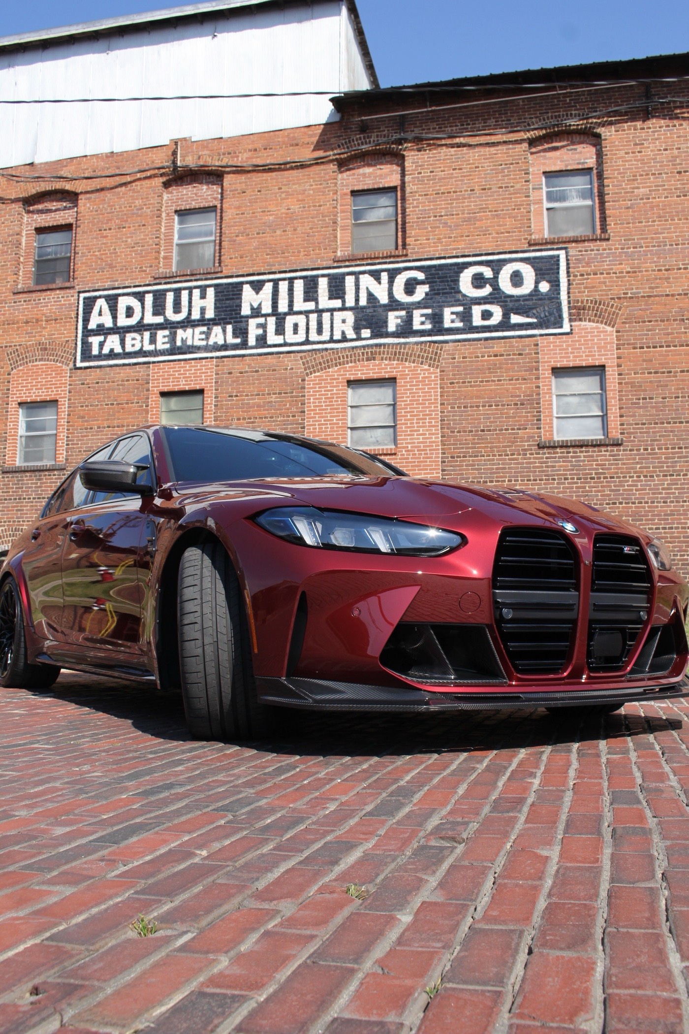 Maroon 2026 BMW M3 Competition parked in front of a brick building